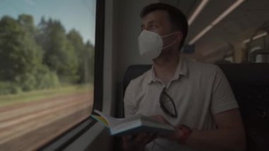 Male passenger in mask sits in passenger seat on modern train in Germany and reads book during coronavirus pandemic. Male in protective mask reading a book on a railroad trip sitting by the window 