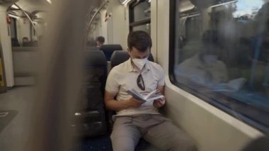 Commuter wearing protective mask reads book on train trip through Germany while sitting by window. A masked train passenger reads book in Bavaria. Travel by train concept. Covid transport fare rules