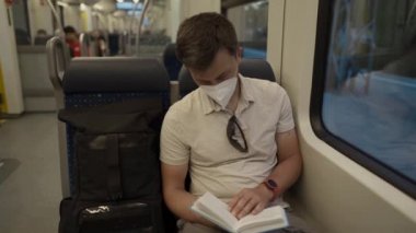 Male passenger in mask sits in passenger seat on modern train in Germany and reads book during coronavirus pandemic. Male in protective mask reading a book on a railroad trip sitting by the window 