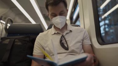 Commuter wearing protective mask reads book on train trip through Germany while sitting by window. A masked train passenger reads book in Bavaria. Travel by train concept. Covid transport fare rules