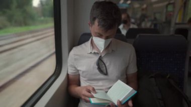 Passenger on public transport is reading a book while sitting by window wearing a protective mask. A man traveling on a Deutsche Trains wearing a protective mask and reading a book. German railroad. 