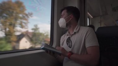 Commuter wearing protective mask reads book on train trip through Germany while sitting by window. A masked train passenger reads book in Bavaria. Travel by train concept. Covid transport fare rules
