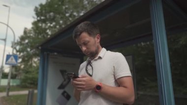 Young caucasian man face beard at the bus stop waiting for the bus in the hands of a mobile phone sends messages. Theme of urban transport and mobility, traveling around Europe in comfortable