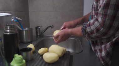 Bachelor is cooking dinner at home in kitchen. Healthy food theme, man peels the skin on raw potato in close-up over kitchen sink. Non GMO vegetables from the farm. Mans hands peeling potato peels. 