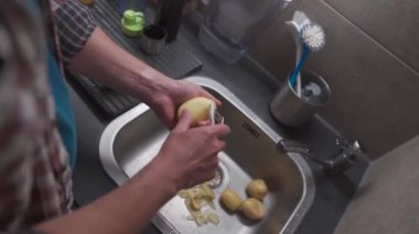 Bachelor is cooking dinner at home in kitchen. Healthy food theme, man peels the skin on raw potato in close-up over kitchen sink. Non GMO vegetables from the farm. Mans hands peeling potato peels. 
