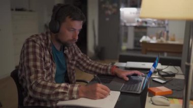 Male American student studies online at home in studio apartment sitting at desk with laptop and taking notes in workbook. Student uses earphones to write notes, watches video online webinar. 