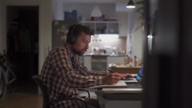 Theme of remote education. American student studying online using a laptop and wireless headphones at home. A man watches an online learning video and takes notes with a pen in a notebook. 