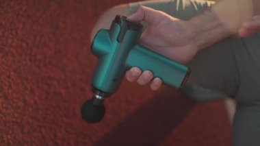 A male athlete massages muscles and tendons with a massage percussion device after a workout at the stadium. Sportsman uses electric massager gun in hand massaging the muscle. Sports recovery concept