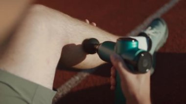 Athletic male massages muscles with hand massage gun, recovering from stadium running workout. . Body treatment with handheld wireless professional vibration shock massager after training session