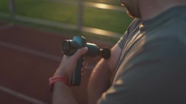 Athletic male massages muscles with hand massage gun, recovering from stadium running workout. . Body treatment with handheld wireless professional vibration shock massager after training session