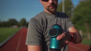 A male athlete massages muscles and tendons with a massage percussion device after a workout at the stadium. Sportsman uses electric massager gun in hand massaging the muscle. Sports recovery concept