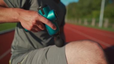 A male athlete massages muscles and tendons with a massage percussion device after a workout at the stadium. Sportsman uses electric massager gun in hand massaging the muscle. Sports recovery concept