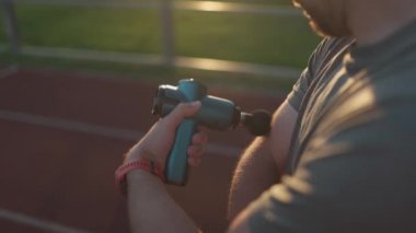 A male athlete massages muscles and tendons with a massage percussion device after a workout at the stadium. Sportsman uses electric massager gun in hand massaging the muscle. Sports recovery concept