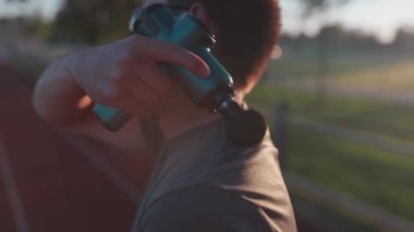 Athletic male massages muscles with hand massage gun, recovering from stadium running workout. . Body treatment with handheld wireless professional vibration shock massager after training session