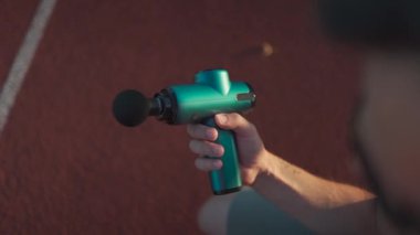 Athletic male massages muscles with hand massage gun, recovering from stadium running workout. . Body treatment with handheld wireless professional vibration shock massager after training session