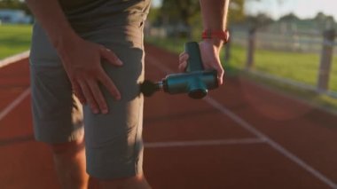 A male athlete massages muscles and tendons with a massage percussion device after a workout at the stadium. Sportsman uses electric massager gun in hand massaging the muscle. Sports recovery concept
