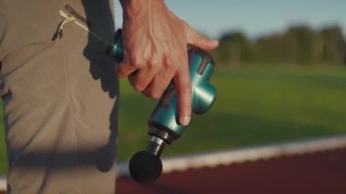 A male athlete massages muscles and tendons with a massage percussion device after a workout at the stadium. Sportsman uses electric massager gun in hand massaging the muscle. Sports recovery concept