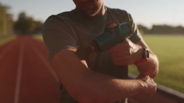 Athletic male massages muscles with hand massage gun, recovering from stadium running workout. . Body treatment with handheld wireless professional vibration shock massager after training session