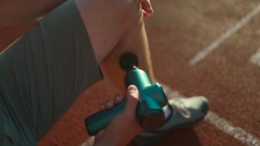 A male athlete massages muscles and tendons with a massage percussion device after a workout at the stadium. Sportsman uses electric massager gun in hand massaging the muscle. Sports recovery concept
