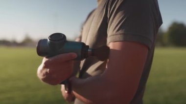 A male athlete massages muscles and tendons with a massage percussion device after a workout at the stadium. Sportsman uses electric massager gun in hand massaging the muscle. Sports recovery concept