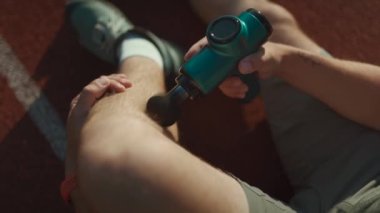 A male athlete massages muscles and tendons with a massage percussion device after a workout at the stadium. Sportsman uses electric massager gun in hand massaging the muscle. Sports recovery concept