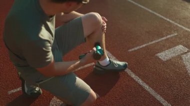 Athletic male massages muscles with hand massage gun, recovering from stadium running workout. . Body treatment with handheld wireless professional vibration shock massager after training session