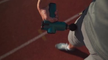A male athlete massages muscles and tendons with a massage percussion device after a workout at the stadium. Sportsman uses electric massager gun in hand massaging the muscle. Sports recovery concept