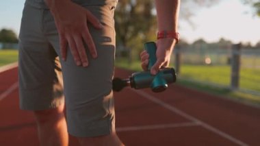 Athletic male massages muscles with hand massage gun, recovering from stadium running workout. . Body treatment with handheld wireless professional vibration shock massager after training session