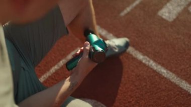 Athletic male massages muscles with hand massage gun, recovering from stadium running workout. . Body treatment with handheld wireless professional vibration shock massager after training session