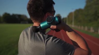 A male athlete massages muscles and tendons with a massage percussion device after a workout at the stadium. Sportsman uses electric massager gun in hand massaging the muscle. Sports recovery concept