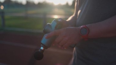 Athletic male massages muscles with hand massage gun, recovering from stadium running workout. . Body treatment with handheld wireless professional vibration shock massager after training session
