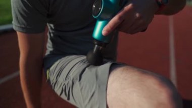 Athletic male massages muscles with hand massage gun, recovering from stadium running workout. . Body treatment with handheld wireless professional vibration shock massager after training session
