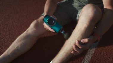 A male athlete massages muscles and tendons with a massage percussion device after a workout at the stadium. Sportsman uses electric massager gun in hand massaging the muscle. Sports recovery concept