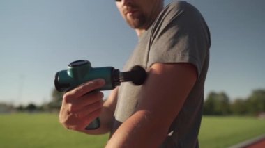 Athletic male massages muscles with hand massage gun, recovering from stadium running workout. . Body treatment with handheld wireless professional vibration shock massager after training session