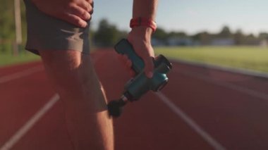 Athletic male massages muscles with hand massage gun, recovering from stadium running workout. . Body treatment with handheld wireless professional vibration shock massager after training session