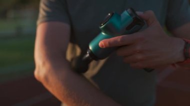 A male athlete massages muscles and tendons with a massage percussion device after a workout at the stadium. Sportsman uses electric massager gun in hand massaging the muscle. Sports recovery concept