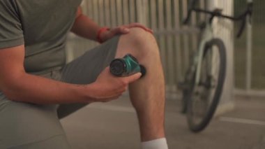 Male cyclist massages muscles and tendons with massage percussion device after cycling workout. Athlete uses an electric pistol massager in his hand, massaging the muscles. Sports recovery concept. 