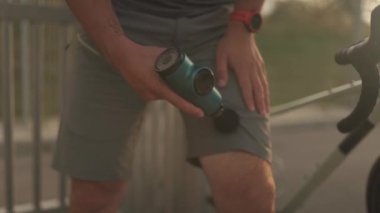 Male cyclist massages muscles and tendons with massage percussion device after cycling workout. Athlete uses an electric pistol massager in his hand, massaging the muscles. Sports recovery concept. 
