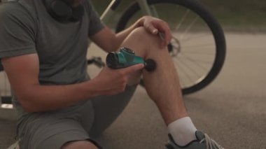 Cyclist athlete massages body with percussion therapy gun to relieve post workout muscle aches and pains on cycling workout. Massage percussion device after workout. Warming up muscles with massager