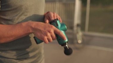 Male cyclist massages muscles and tendons with massage percussion device after cycling workout. Athlete uses an electric pistol massager in his hand, massaging the muscles. Sports recovery concept. 