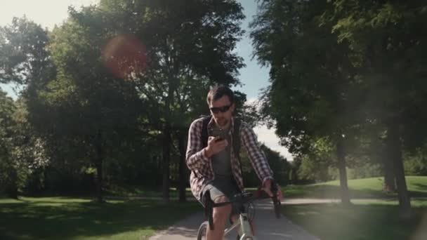 在阳光充足的天气里 一个骑自行车背着背包的男人骑自行车在公园里 一边骑自行车一边打电话 一位骑自行车的人在公园里骑自行车 拿着背包在电话里交谈 — 图库视频影像