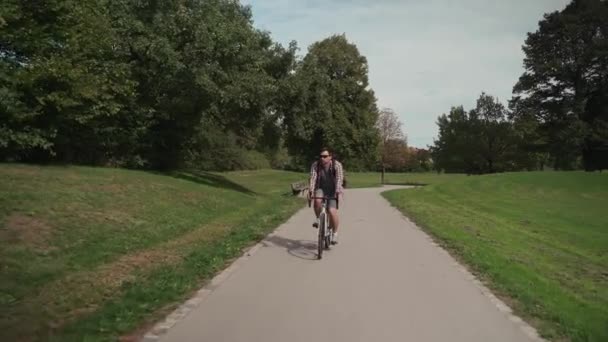 シャツを着た男と ドイツのミュンヘンのシティガーデンのサイクルパス上のリュックサック自転車に乗っています 公園の自転車道で学生サイクリストサイクリング 緑のエリアで街のサイクルに乗るバックパックを持つ男性 — ストック動画