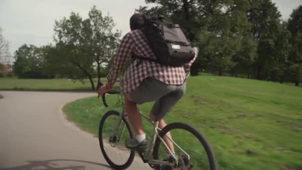 シャツを着た男と ドイツのミュンヘンのシティガーデンのサイクルパス上のリュックサック自転車に乗っています 公園の自転車道で学生サイクリストサイクリング 緑のエリアで街のサイクルに乗るバックパックを持つ男性 — ストック動画