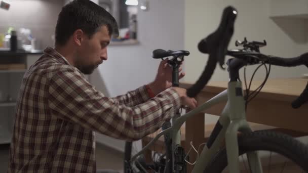 若い男が自宅のスタジオアパートに自転車のアクセサリーをインストールします 男は家で自転車をセットアップします 自宅の砂利の自転車で男性の固定自転車 新しい季節のための自転車の修理と準備の概念 — ストック動画