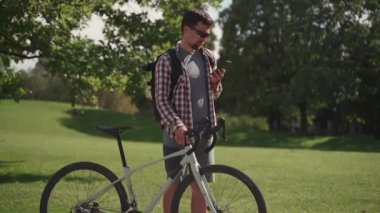 Bisikletinin yanında yürüyen bir bisikletçi güneşli bir havada parkta internette sohbet ediyor. Bisikletli ve akıllı telefonlu bir yolcu. Adam yaz yürüyüşünde bisikletli telefondan hoşlanıyor.. 