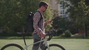 Bir erkek bisikletinin yanında yürür ve Almanya 'da güneşli havada akıllı telefon kullanır. Adam bisikletini itiyor ve internette sörf yapıyor. Bisiklet sürerken sohbet etmek. Modern yaşam tarzı, bağlantı. 