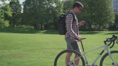 Bisikletinin yanında yürüyen bir bisikletçi güneşli bir havada parkta internette sohbet ediyor. Bisikletli ve akıllı telefonlu bir yolcu. Adam yaz yürüyüşünde bisikletli telefondan hoşlanıyor.. 
