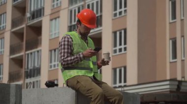 İnşaatçı şantiyede inşaat elemanlarının üzerine oturup kahve içerek ve akıllı telefondan internette sörf yaparak rahatlıyor. Miğferli mimar iş yerinde dinleniyor ve kahve içiyor.. 