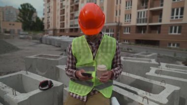 İnşaat işçisi inşaat alanında dinleniyor kahve içiyor ve iş yerinde otururken akıllı telefon kullanıyor. İş yerindeki kahve molasında mimar sohbet ediyor ve akıllı telefon kullanıyor.. 