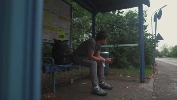 疲れとストレスのある男がバス停に座って 時計や道路を見て 輸送のための長い時間を待ちます 乗客はミュンヘンのドイツ駅で電話を使用しています バスの待ち時間が長くなる — ストック動画